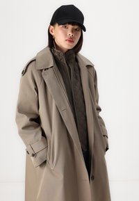 Abrigo beige tipo trench con un amplio cuello, acentos de botones y bolsillos laterales, llevado sobre una chaqueta interna acolchada marrón. Lleva puesta una gorra negra.