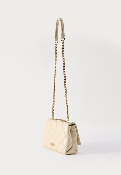 Μπεζ διακοσμημένη τσάντα ώμου με χρυσή αλυσίδα ως λουρί και μικρή χρυσή πλακέτα με το λογότυπο της μάρκας στο μπροστινό μέρος, απεικονίζεται κρεμασμένη σε απλό φόντο.