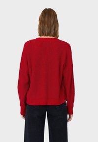Pull en tricot rouge à coupe ample, épaules tombantes et texture côtelée. L'ourlet arrive à la taille, porté avec un pantalon sombre.
