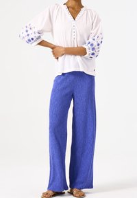 Vit skjortblus med blått blommönster på ärmarna, kombinerad med strukturerade blå vida byxor. Sandaler fullbordar outfiten.