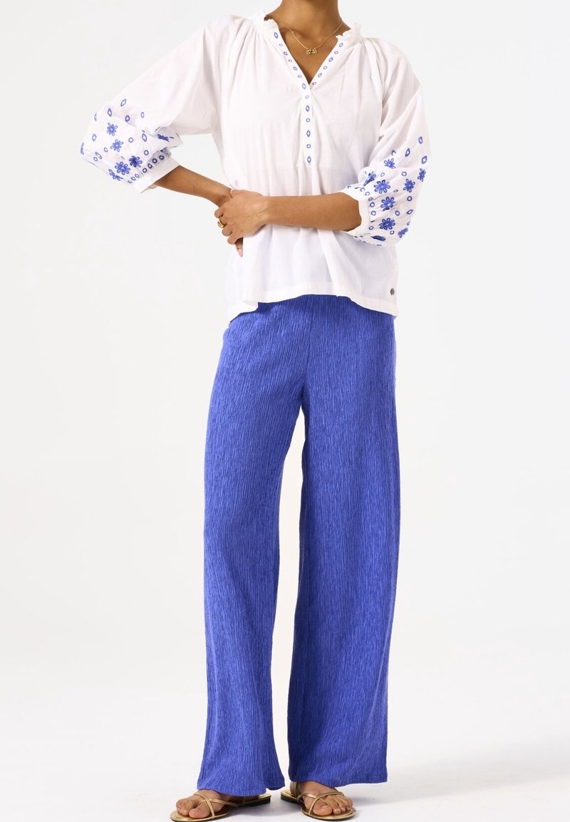 Vit skjortblus med blått blommönster på ärmarna, kombinerad med strukturerade blå vida byxor. Sandaler fullbordar outfiten.