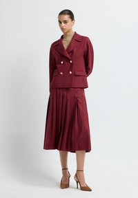 Woman wearing a maroon double-breasted blazer with gold buttons, matching pleated midi skirt, tan turtleneck, and brown high-heeled shoes.