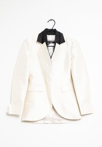 Bílý saténový blazer s černým límcem, jednořadým zapínáním, dvěma předními kapsami a dlouhými rukávy visící na černém ramínku na bílém pozadí.