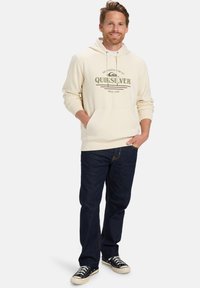 Sudadera crema con bolsillo frontal, cordones y texto gráfico verde. Combinada con jeans azul oscuro y zapatillas negras.