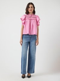Femme debout portant un haut à volants rose à manches courtes et un jean large bleu, avec des chaussures noires pointues sur un fond uni.