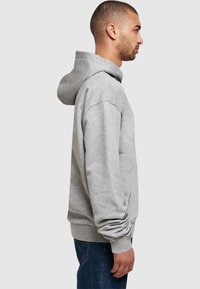 Sweatshirt à capuche grise en tissu doux, avec une poche avant, des poignets côtelés et un ourlet côtelé. Présenté de profil sur un fond neutre.
