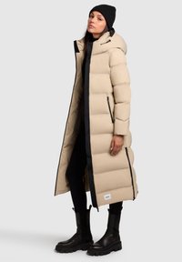 Doudoune beige avec des bordures noires, dotée d'une capuche, d'un design matelassé vertical et de poches zippées. Portée sur des vêtements et des bottes noires.