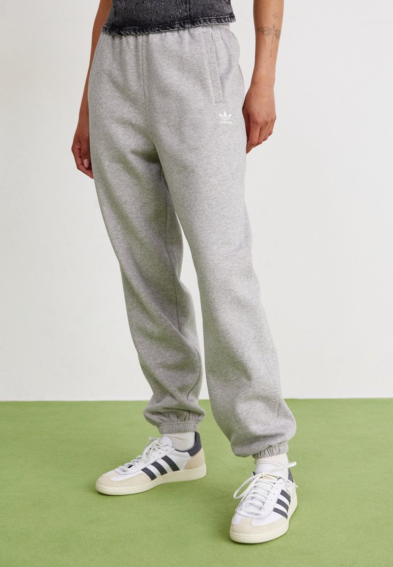 Pantalon de survêtement gris en mélange de coton, avec une taille élastique, des chevilles légèrement resserrées et un logo blanc. Porté avec des sneakers blancs et noirs.