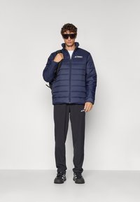 Chaqueta acolchada de marina con costuras verticales, cierre de cremallera y cuello alto; combinada con pantalones oscuros y zapatillas negras. Logo en el pecho.