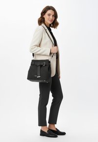 Mujer con blazer beige, blusa negra y pantalones negros, sosteniendo un bolso estructurado negro con detalle de cinturón en el hombro.