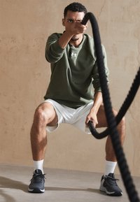Grüner Sportpullover, weiße Shorts, schwarze Trainingsschuhe mit strukturiertem Muster, weiße Socken. Hält ein dickes, schwarzes Battle Rope.