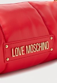 Raudona dviguba odinė rankinė su auksiniu "LOVE MOSCHINO" logotipu ir auksiniais smeigėmis. Lygus paviršius su struktūrizuotu, stačiakampiu dizainu.