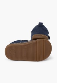 Pantofi pentru copii, de culoare bleu marin, cu un superior texturat, curea cu arici și talpă din cauciuc maro cu un model ondulat și amprenta logo-ului.