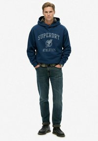 Marineblauwe hoodie met de tekst "SUPERDRY ATHLETICS", gecombineerd met donkere spijkerbroek en zwarte leren schoenen. Duidelijk geruit patroon zichtbaar bij de manchetten.