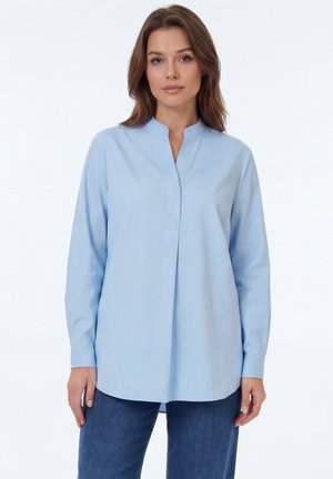 Woman wearing a light blue long-sleeve blouse with a stand collar and dark blue jeans, standing against a white background.