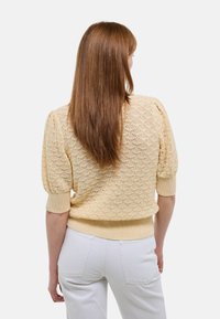 Un pull en tricot jaune avec des manches bouffantes, présentant un motif en dentelle et un ourlet côtelé. Associé à un pantalon blanc. Vue de dos.