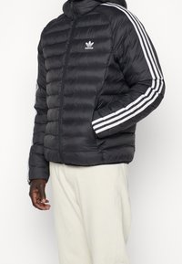 Svart puffjacka med dragkedja fram, huva och vita ränder på ärmarna. Har en broderad logotyp på bröstet och sidofickor.