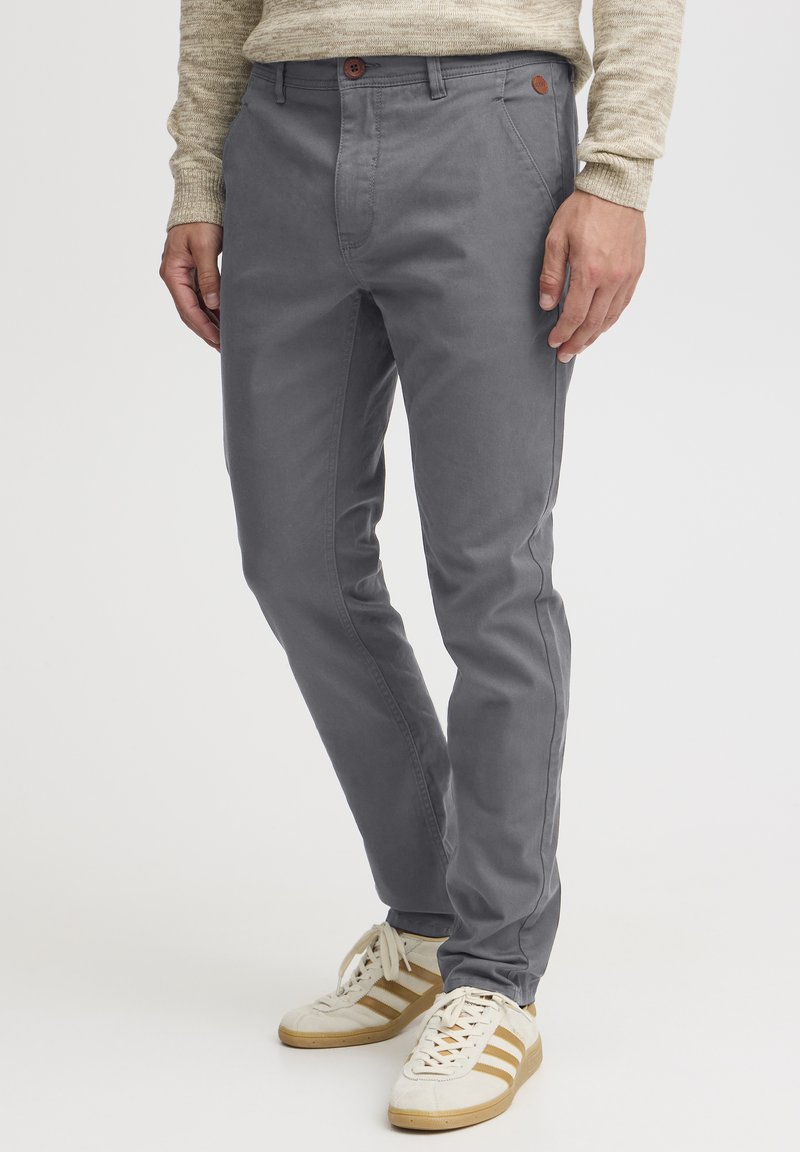 Pantalones grises de corte ajustado, con cierre de botón y bolsillos frontales. Combinados con zapatillas blancas con franjas doradas.