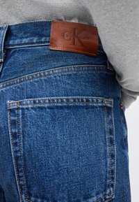 Κοντινό πλάνο πίσω τσέπης μπλε τζιν με καφέ δερμάτινο λογότυπο Calvin Klein και πρόσωπο που φορά γκρι ριγωτό τοπ.