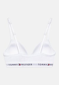 Bralette blanche avec des bretelles réglables et une bande de logo affichant "TOMMY HILFIGER" en noir, rouge et bleu. Texture de tissu lisse.