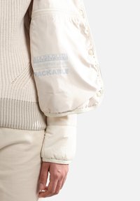 Světle puffy bunda v off-white, se prošívaným designem, měkkou texturou a značkováním na rukávu; sbalitelná s druky.