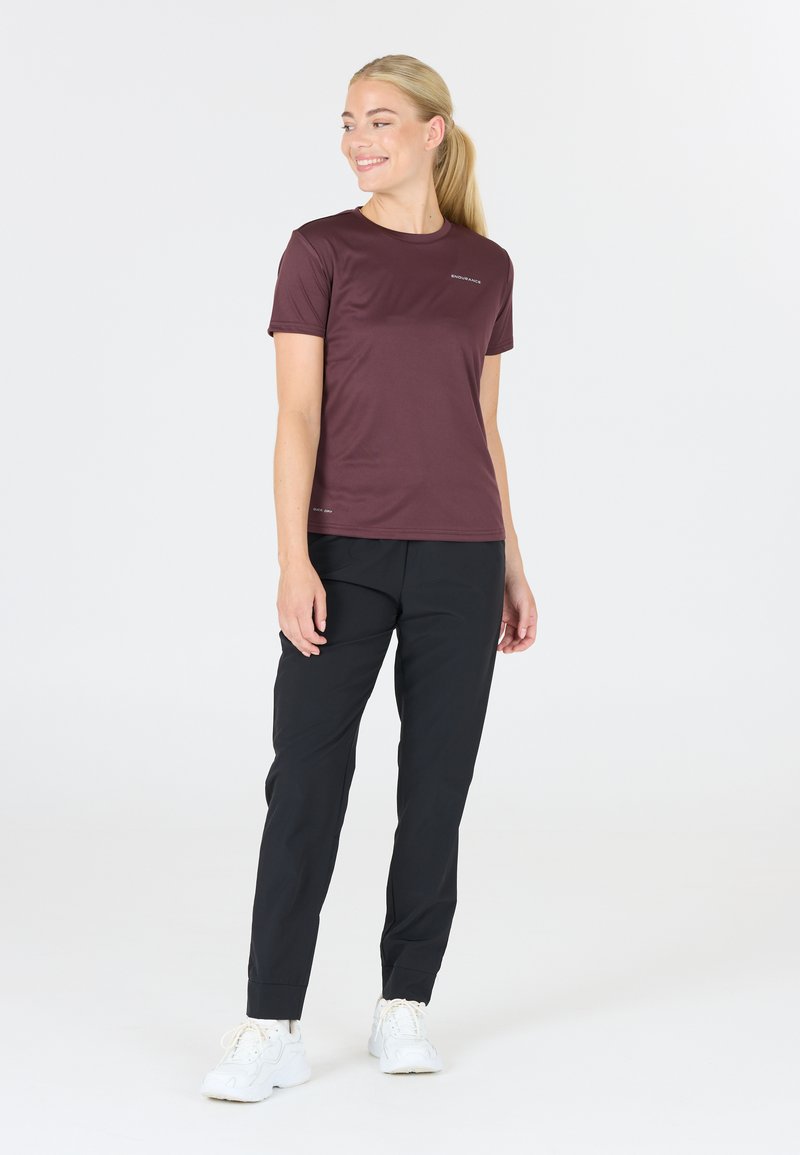 Kastanjefärgad kortärmad träningströja med en slät textur, matchad med svarta tapered byxor. Modellen bär vita träningsskor. Enkel bakgrund.