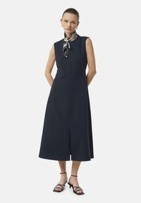 Navy-blaues ärmelloses Kleid mit geradem Schnitt, hoher Halslinie, Schlitz vorne und einem gemusterten Schal am Ausschnitt. Schwarze Sandaletten mit Riemen.
