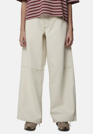 Personne portant une chemise ample à rayures rouges et blanches, un pantalon crème à jambes larges et des baskets marron, debout devant un fond uni.
