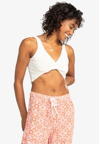 Biely krátky top s prekrúteným predným dizajnom, kombinovaný s oranžovými širokými nohavicami so vzorom, ktoré majú sťahovaciu šnúrku v páse a kvetinové motívy.