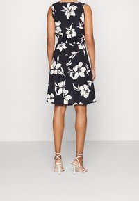Schwarzes Blumenkleid mit weißen Blumen, tailliert, ärmelloses Design. Kombiniert mit weißen Riemchenschuhen, die einen lässigen, aber eleganten Look zeigen.