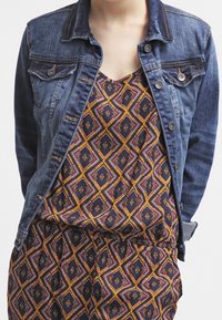 Veste en jean avec devant boutonné, deux poches poitrine, bords usés, portée par-dessus une robe à motifs avec des formes géométriques en violet, jaune et noir.