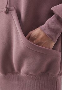 Lila sweatshirt med en kenguruficka, ribbade ärmslut och en slät, mjuk textur. Tyget ser varmt och avslappnat ut.