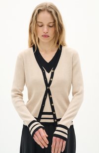 Cardigan beige à côtes avec finition en V noir, comportant des boutons dorés et des poignets rayés noirs. Texture lisse, design ajusté et manches longues.