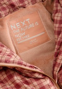 Miękka różowa i czerwona podszewka w kratkę kurtki z zszytą metką z napisem "NEXT THE FUTURE IS NOW EST. 1982," ukazująca szczegóły zamka błyskawicznego.