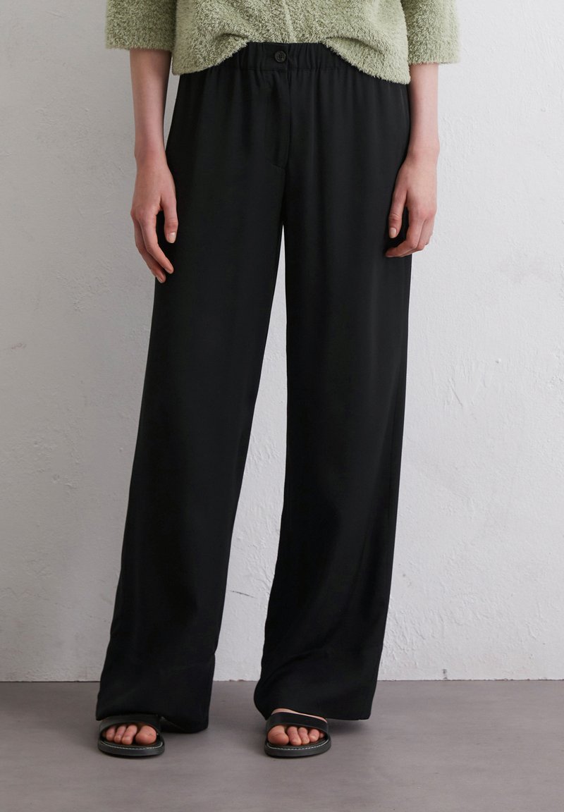 Schwarze weit geschnittene Hosen mit glatter Textur, ausgestattet mit einem elastischen Gummibund und einem Knopfverschluss. Kombiniert mit schwarzen flachen Sandalen.