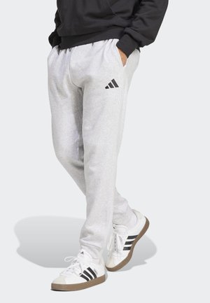 Person trägt hellgraue Adidas-Sweatpants und weiße Adidas-Sneaker mit schwarzen Streifen, steht mit den Händen in den Taschen.