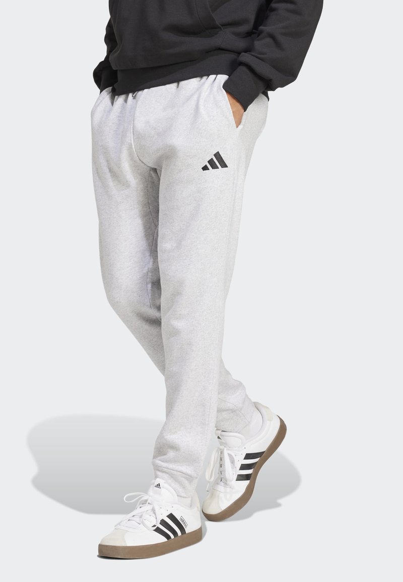 Person trägt hellgraue Adidas-Sweatpants und weiße Adidas-Sneaker mit schwarzen Streifen, steht mit den Händen in den Taschen.