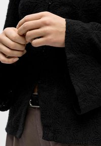 Handen die de manchetten van een zwart gestructureerd overhemd met lange mouwen aanpassen, gedragen met een bruine broek en een zwarte riem met een zilveren gesp.