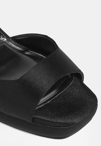 Schwarze Plateau-Sandale mit satinierter Oberseite, Überkreuz-Design, offener Zehenpartie und glatter schwarzer Sohle. Keine sichtbaren Beschläge oder Verzierungen.