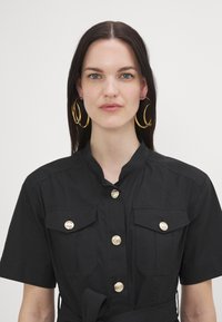 Cămașă utilitară neagră cu mâneci scurte, detalii cu nasturi aurii și două buzunare pe piept. Dispune de un guler mandarin și o talie cu brâu.