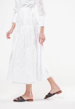 Vrouw in een witte eyelet midi-jurk met lange mouwen en zwarte platte slides die tegen een witte achtergrond loopt.