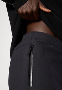 Calças de desporto pretas com um corte slim, apresentando um bolso lateral com fecho e uma textura de tecido suave. A vista em close destaca a cintura.