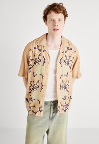 Korte mouw beige shirt met bloemenborduurwerk in marineblauw en mintgroen. Heeft een kraag en knoopsluiting.