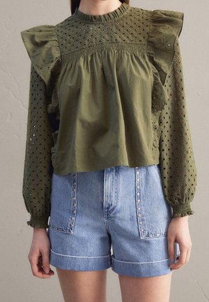 Olivgrüne Bluse mit ruchéärmeln und Lochstickereidetails, kombiniert mit hellblauen Denim-Shorts mit Nietenakzenten und umgeschlagenen Bündchen.