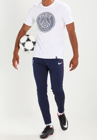 Camiseta blanca con el logo redondo del Paris Saint-Germain, combinada con pantalones deportivos azul marino con franjas laterales rojas, y zapatillas de fútbol negras.