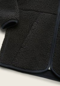 Giacca in pile nera caratterizzata da un tessuto testurizzato, bordi curvi e dettagli in piping scuro a contrasto lungo la cerniera e le tasche.