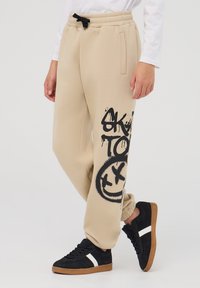 Beżowe dresy z bocznymi kieszeniami i czarnym nadrukiem w stylu graffiti. Elastyczny pas ze sznurkiem, ściągacze przy nogawkach, w połączeniu z czarnymi trampkami.