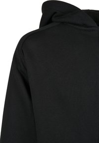 Détail du tissu d'un sweat à capuche noir montrant la couture de l'épaule et le bord de la capuche sur fond blanc uni.