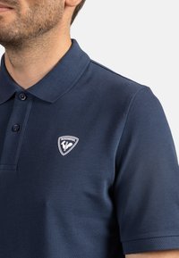 Námořnická modrá polo tričko vyrobené z texturované tkaniny, s límečkem, dvěma knoflíky a bílým logem Rossignol na levé straně hrudníku.