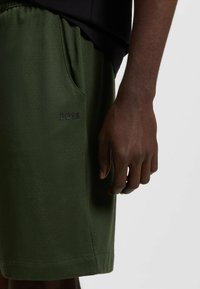 Gröna träningsshorts med en mjuk textur, med perforerade detaljer och en logotyp på benet. Sidofickan är synlig.